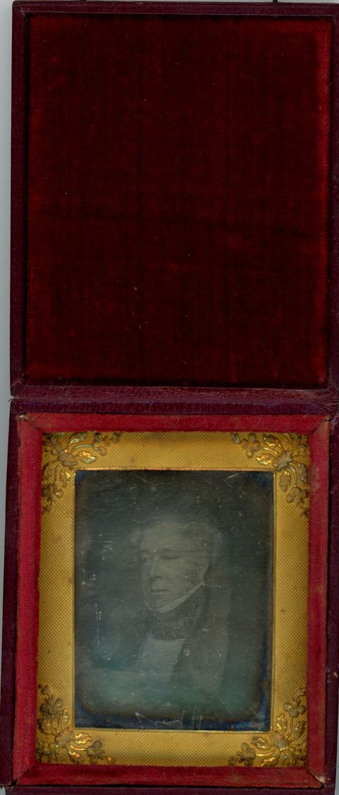 Daguerreotype in an open case. The ghostly image of a clean-shaven man with white or grey hair looks off to our left. The rectangular mat has leafy designs at the corners. The lid of the case is flipped up, the inside lined with dark red velvet.