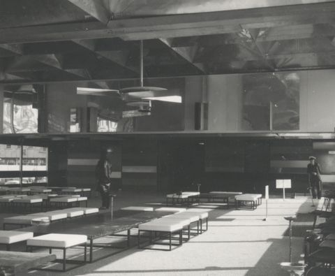 Installation photograph. Black and white photograph of a two-storey-high open space. An abstract sculpture made of discs and strips of metal hangs from the ceiling. On the far wall, the lower level is filled with horizontal bands in different shades. Reflective surfaces are above, on the second level of the open space.